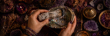 How to smudge your space – hands holding a lit white sage smudge wand above an abalone shell during a home cleansing ritual on a mystical esoteric background