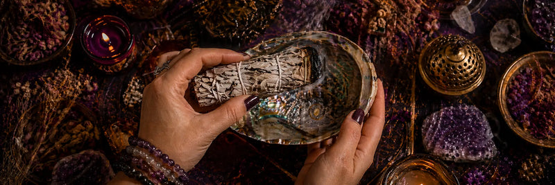 How to smudge your space – hands holding a lit white sage smudge wand above an abalone shell during a home cleansing ritual on a mystical esoteric background